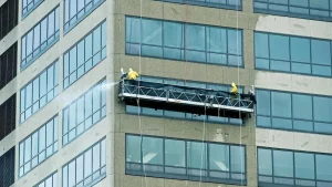 Two window cleaners on a hanging platform pressure wash windows and stone facade of a high rise office building