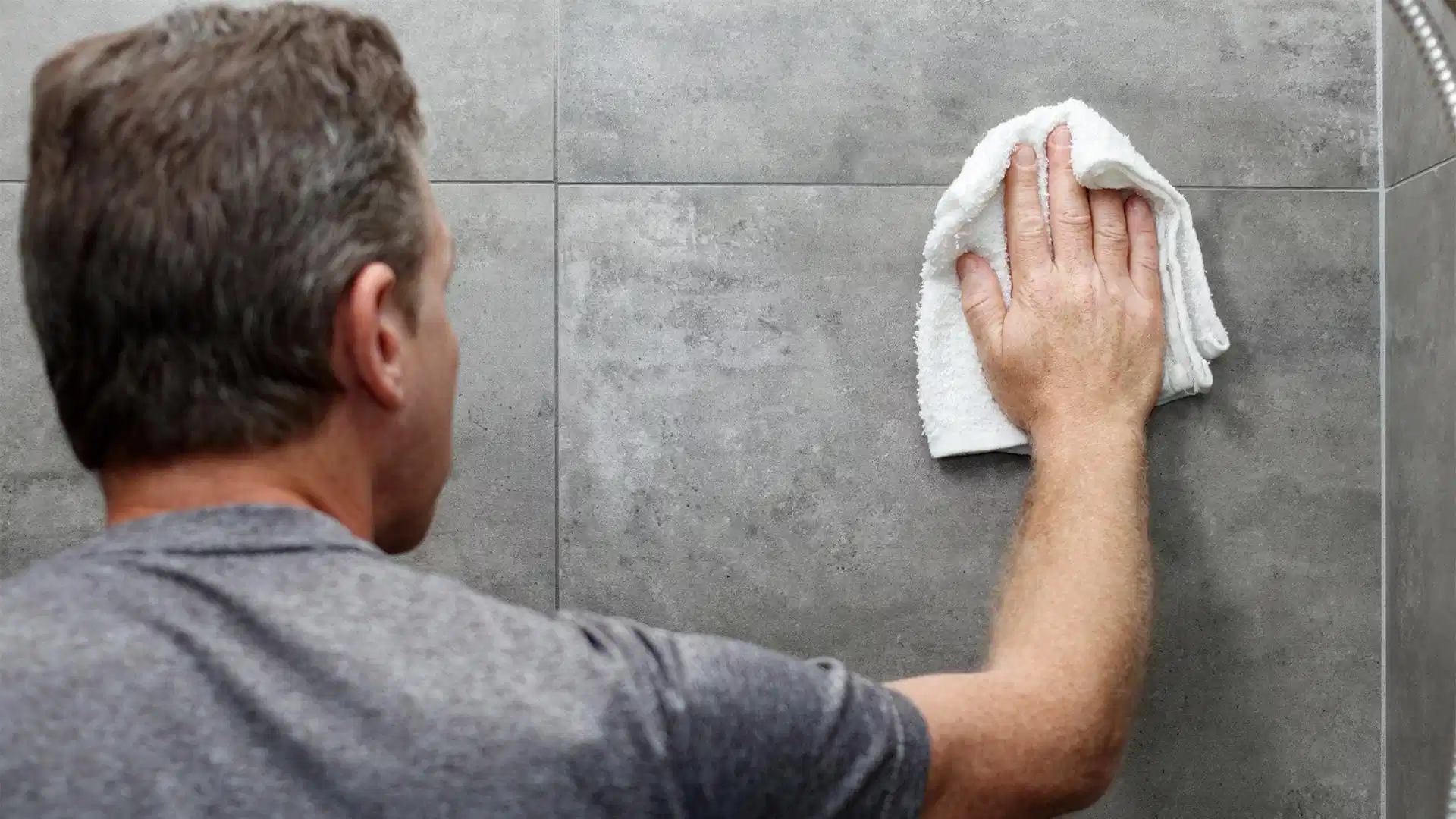 Man scrubs grout lines on grey tiled wall with a white towel